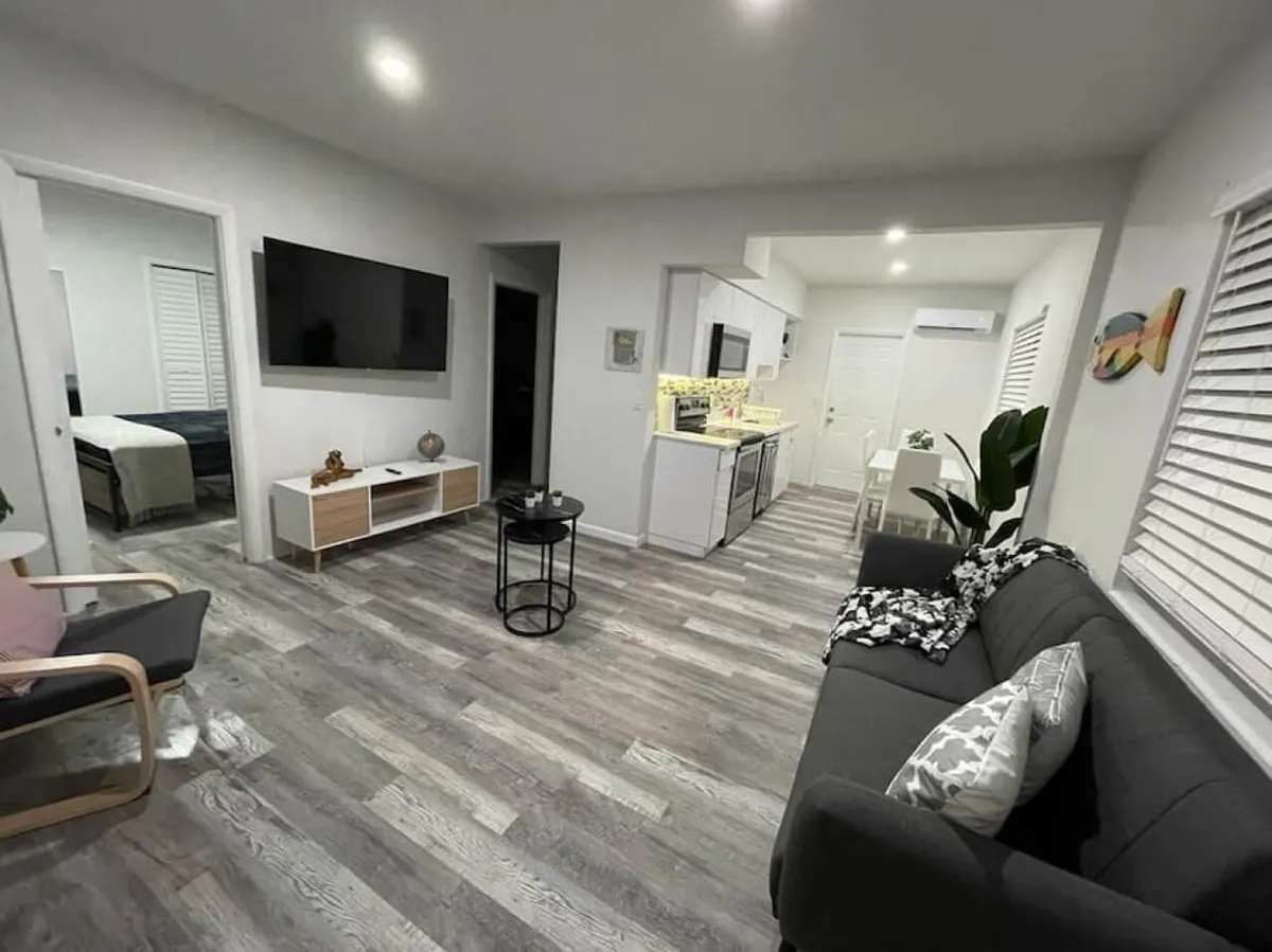 The image showcases a bright, open-plan living space featuring a cozy gray sofa with patterned pillows, a wall-mounted flat screen TV, a modern kitchen with sleek white cabinets and appliances, and a stylish wooden armchair. The space feels airy and inviting, perfect for relaxing after a day in Miami.