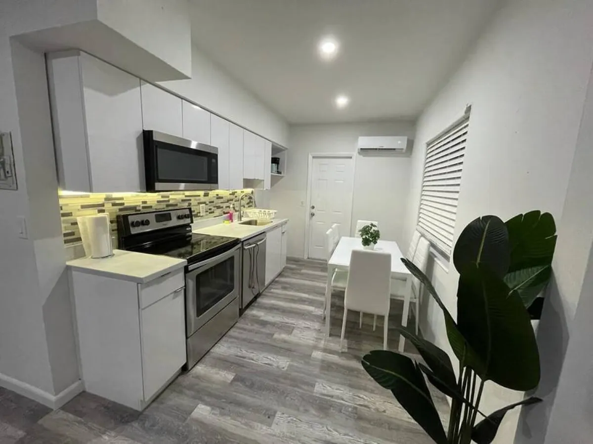 This image showcases a bright, modern kitchen featuring a stainless steel stove and microwave, white cabinetry with under-cabinet lighting, a small white dining table with chairs, and a large green plant in the corner. The space feels open and inviting, perfect for gatherings and casual meals.
