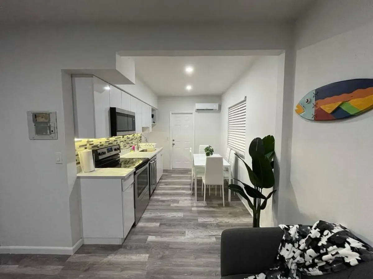 This photo showcases a modern kitchen with white cabinetry and stainless steel appliances, a dining area with white chairs and table, and a gray sofa with a patterned blanket. The colorful fish wall art adds a playful accent. The space is bright and open, ideal for smooth arrivals and daily routines.