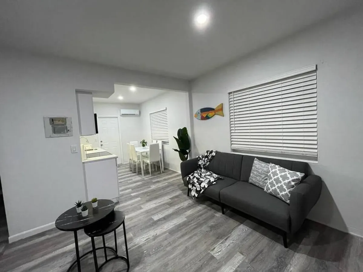 This image displays a calm and inviting living area featuring a dark grey sofa with patterned cushions, small round black nested tables with potted plants, and a white dining table with chairs in the background. The colorful fish wall decoration adds a cheerful touch, and the space feels open, bright, and accessible.