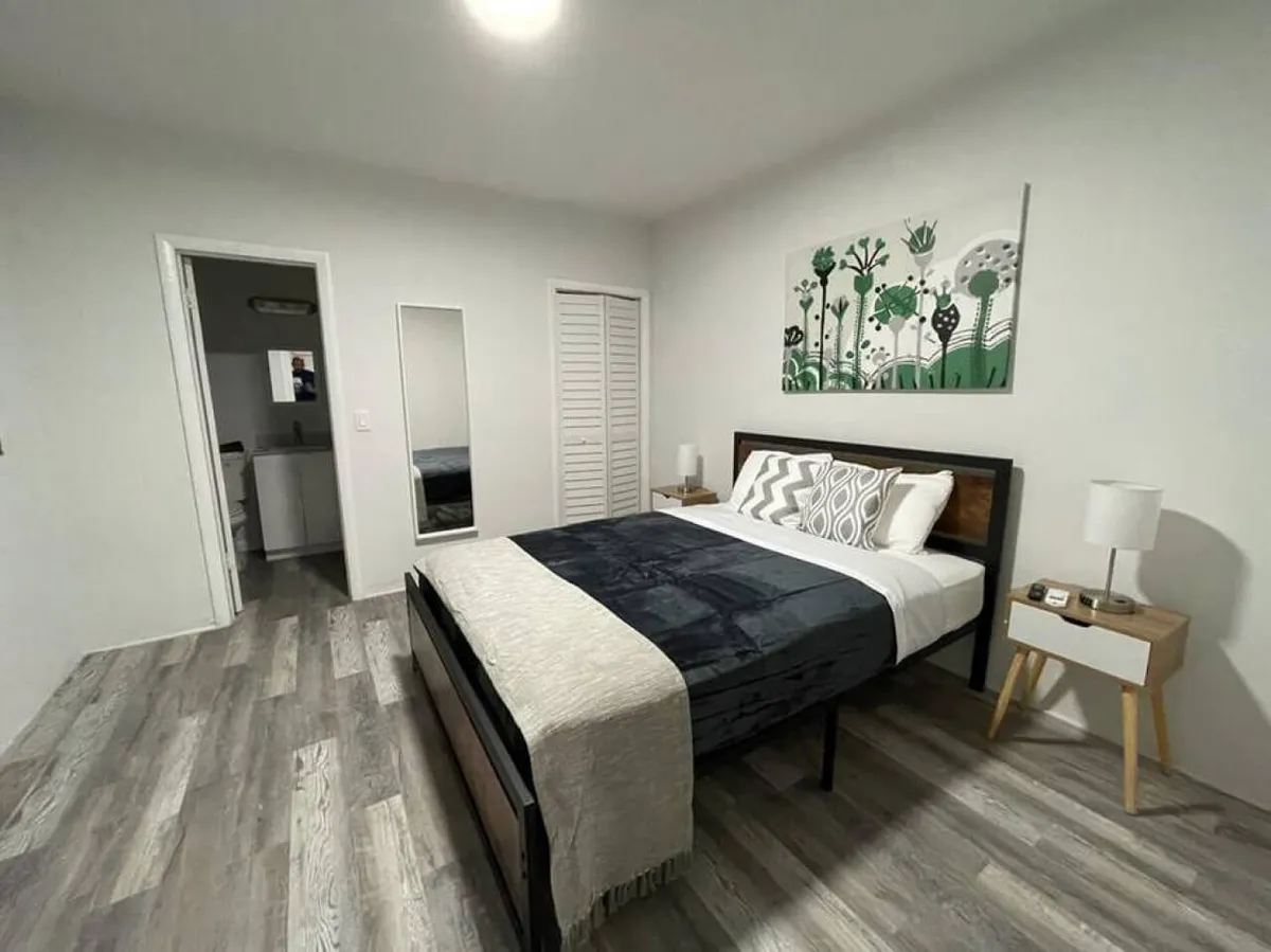A cozy, modern primary bedroom featuring a queen-size bed with stylish patterned pillows and dark bedding, flanked by two wooden nightstands with lamps. The room is brightened by abstract green and white wall art, creating a welcoming and restful ambiance.