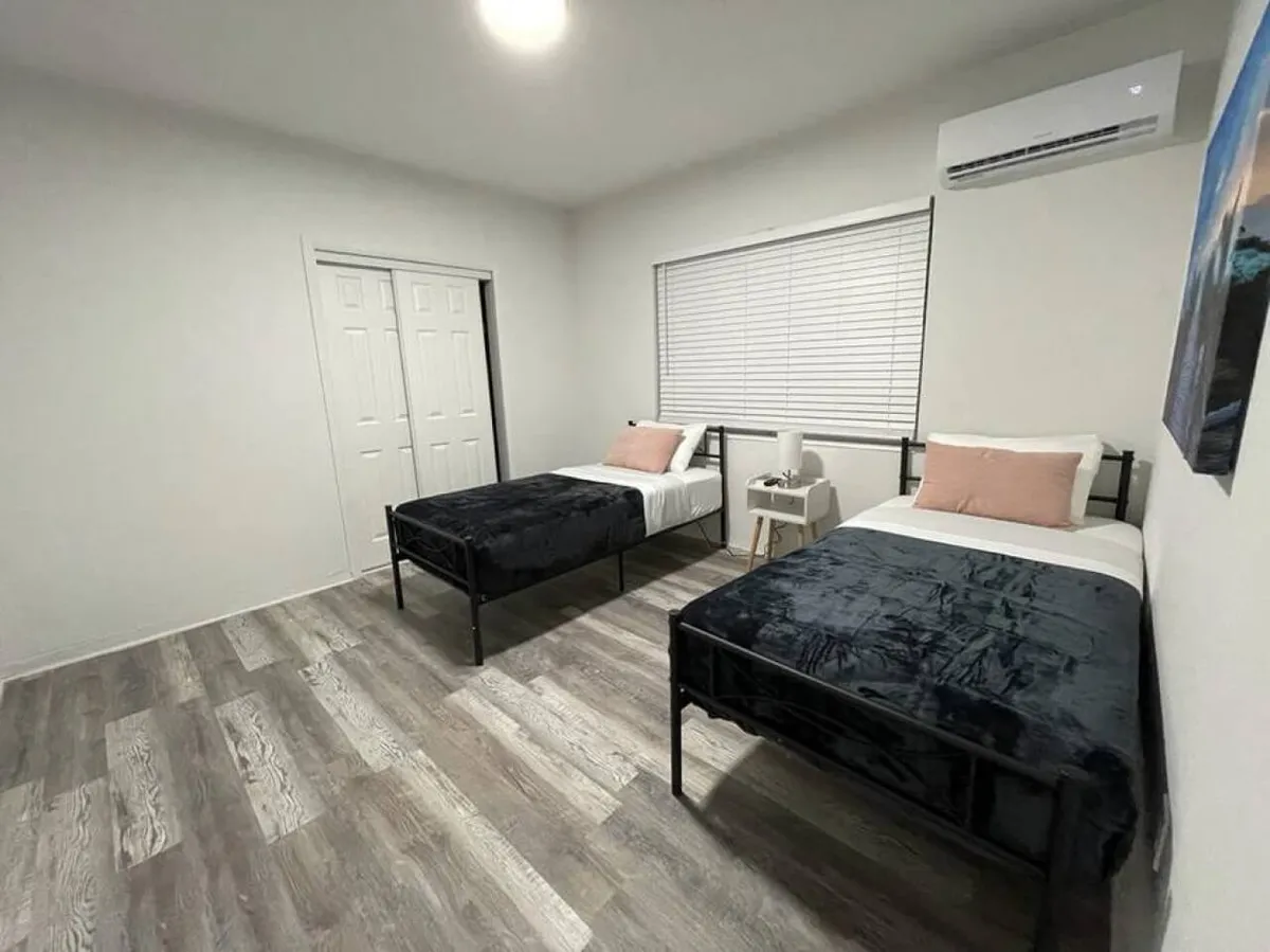 A quiet bedroom with two single beds featuring black metal frames, a white bedside table with a lamp, and a window with closed blinds. The space is climate-controlled with a wall-mounted air conditioning unit, making it ideal for kids, friends, or business travelers.