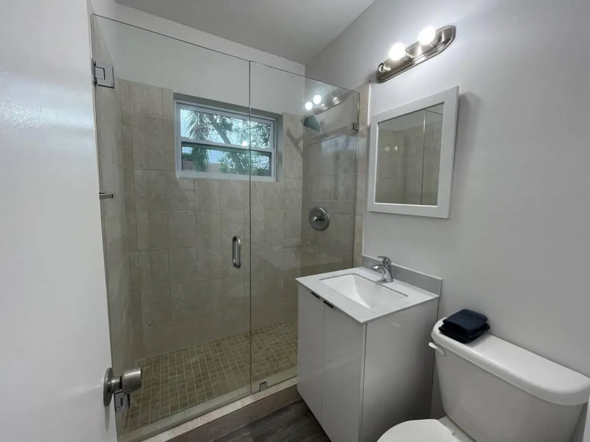 A clean, modern bathroom featuring a glass-enclosed shower lined with beige tiles, a compact white vanity with a mirror, and a toilet topped with neatly folded towels. Soft natural light enters through a small window, enhancing the fresh ambiance.